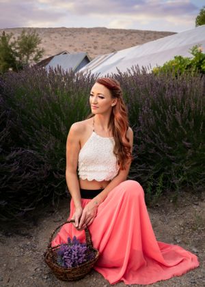 Woman in white lace crop top and pink maxi skirt sits among purple lavender fields holding a basket.