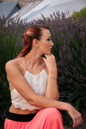 Redhead woman in white lace cami and pink pants sits pensively in front of blooming lavender bushes.