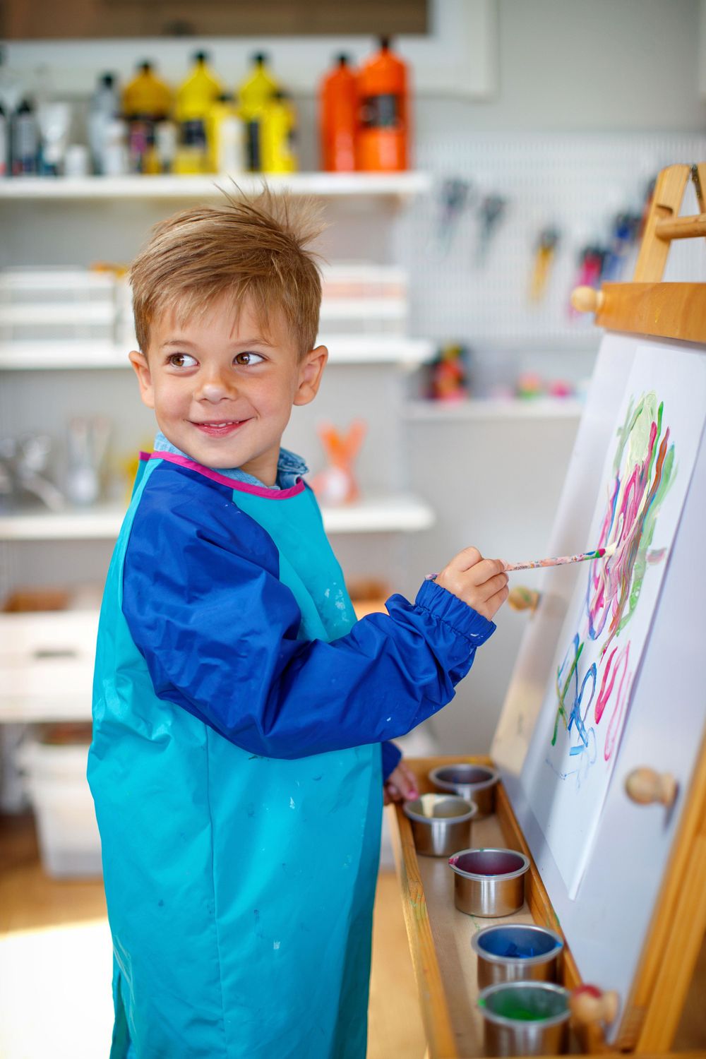 Young child wearing blue art smock painting at an easel with colorful paints.