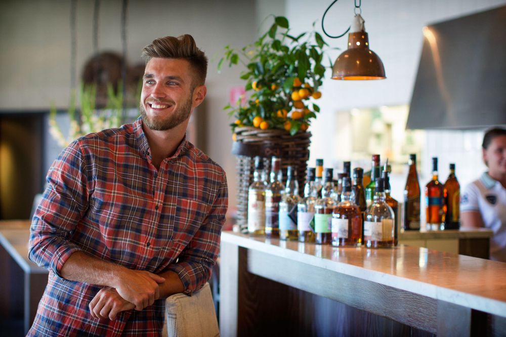 Casual bartender in plaid shirt standing behind bar counter with bottles displayed.