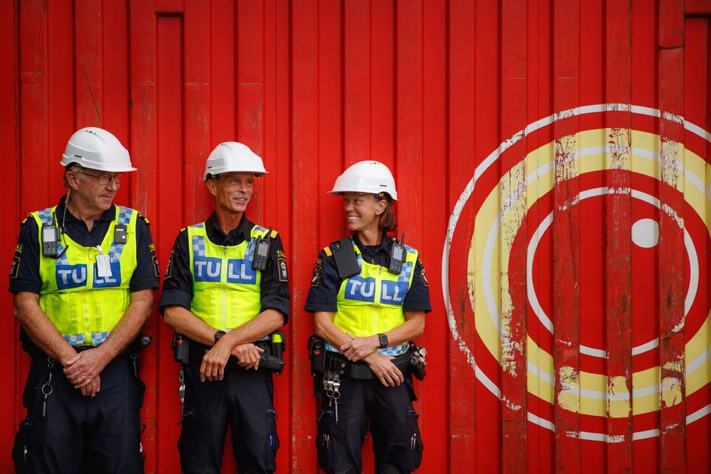 Three security officers in high-vis vests and hard hats standing against a red wall with circular logo.