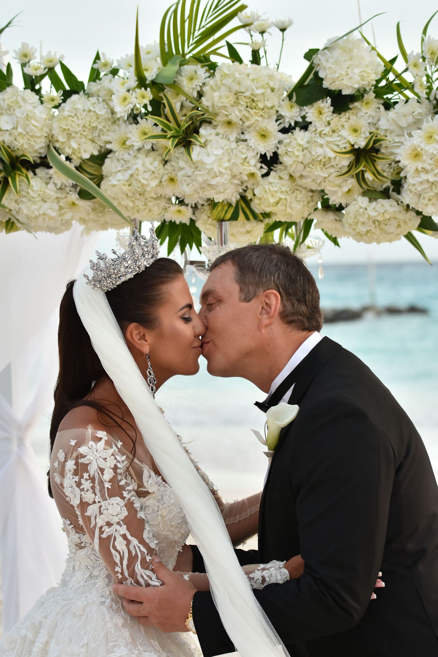Wedding ceremony scene under white floral arch with hydrangeas and palm leaves against ocean backdrop.