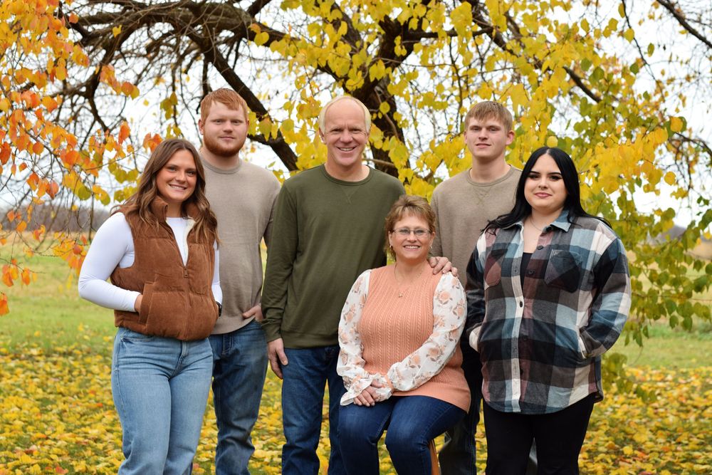 Family poses together in casual attire against a backdrop of vibrant autumn trees with golden leaves.
