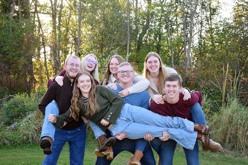 Playful family photo with group members carrying each other outdoors in natural setting.