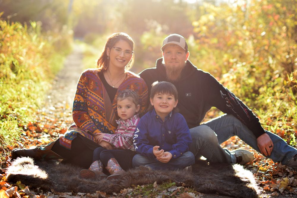 Family sitting together on fallen leaves in autumn sunlight with golden foliage in background.
