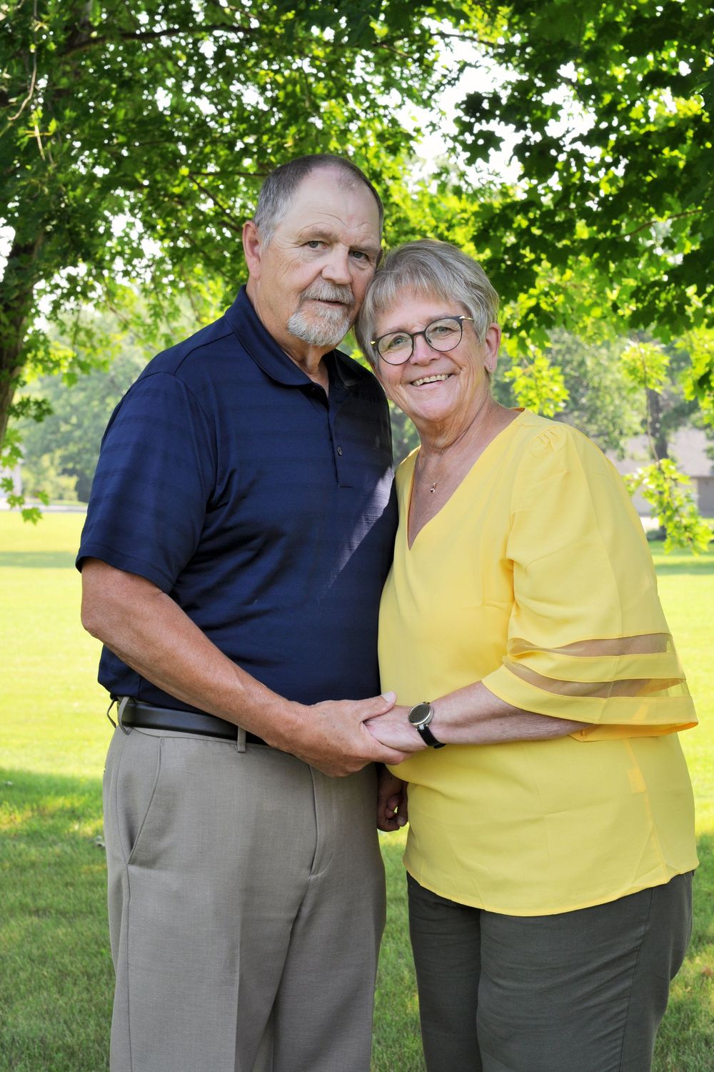 Couple wearing yellow and navy outfits embrace while standing in a sunny green park setting.