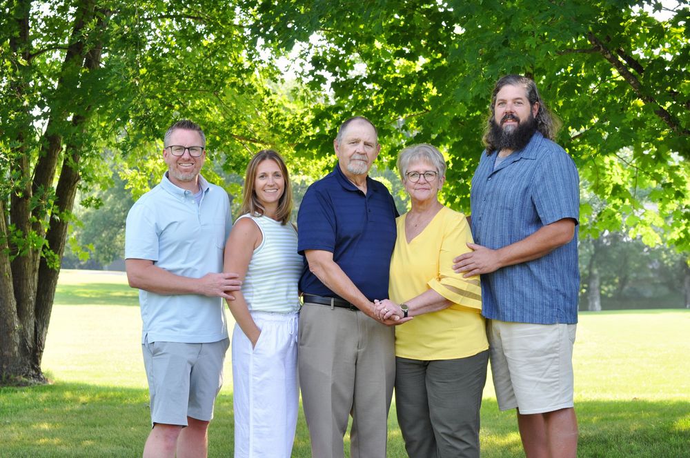 Family members pose together in casual summer attire under green trees in park setting.