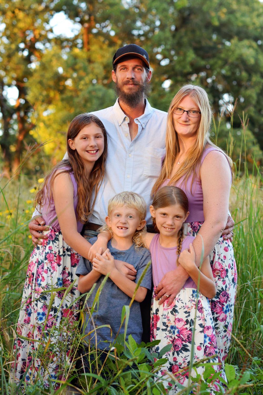Family portrait outdoors with floral dresses and casual attire against a natural summer backdrop.