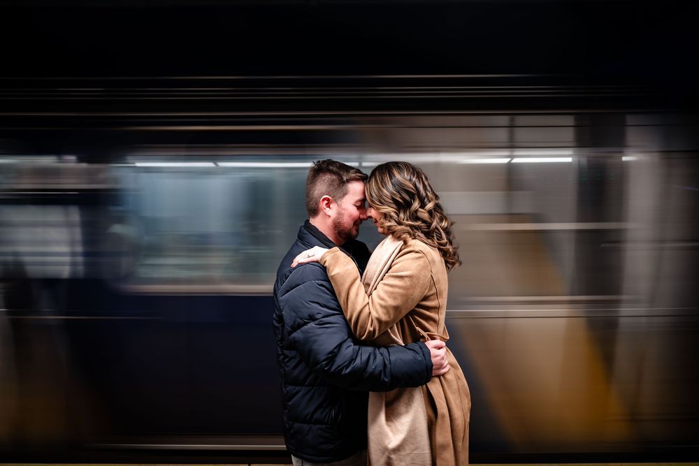 A couple shares an intimate kiss on a train platform as a blurred subway train passes by in the background.