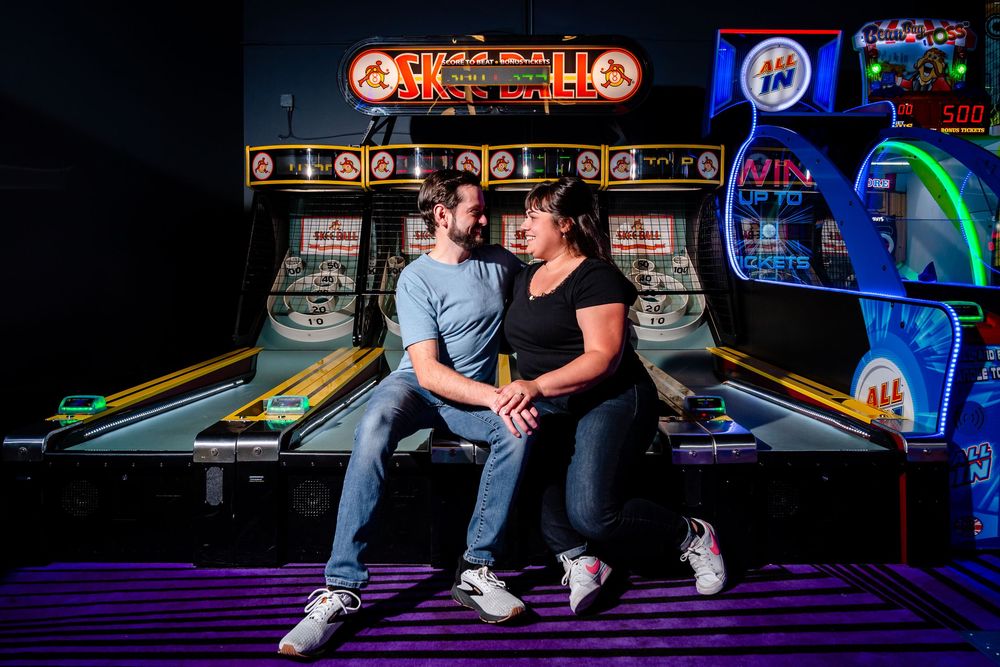 People playing arcade games and skee ball in a dimly lit arcade with neon lighting and colorful game displays.