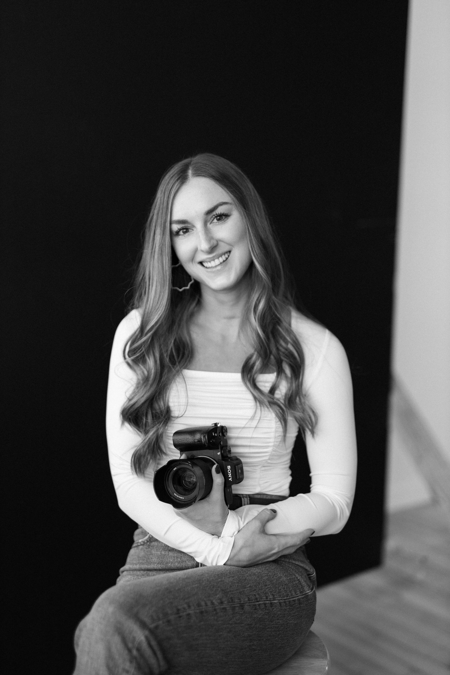 Professional photographer in white sweater holding camera against dark background.