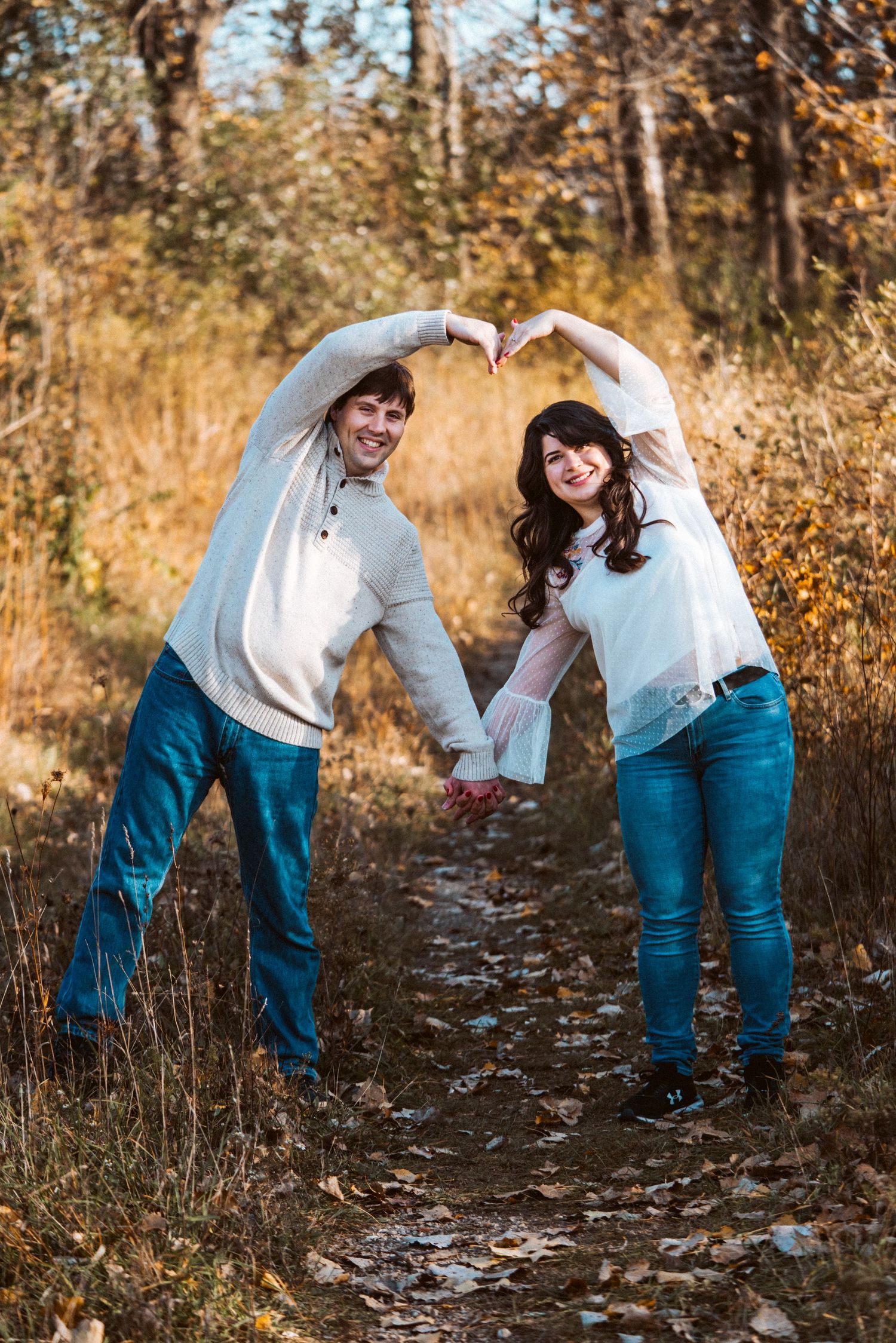 Playful autumn photo session showing couple in matching outfits walking through fallen leaves on wooded path.