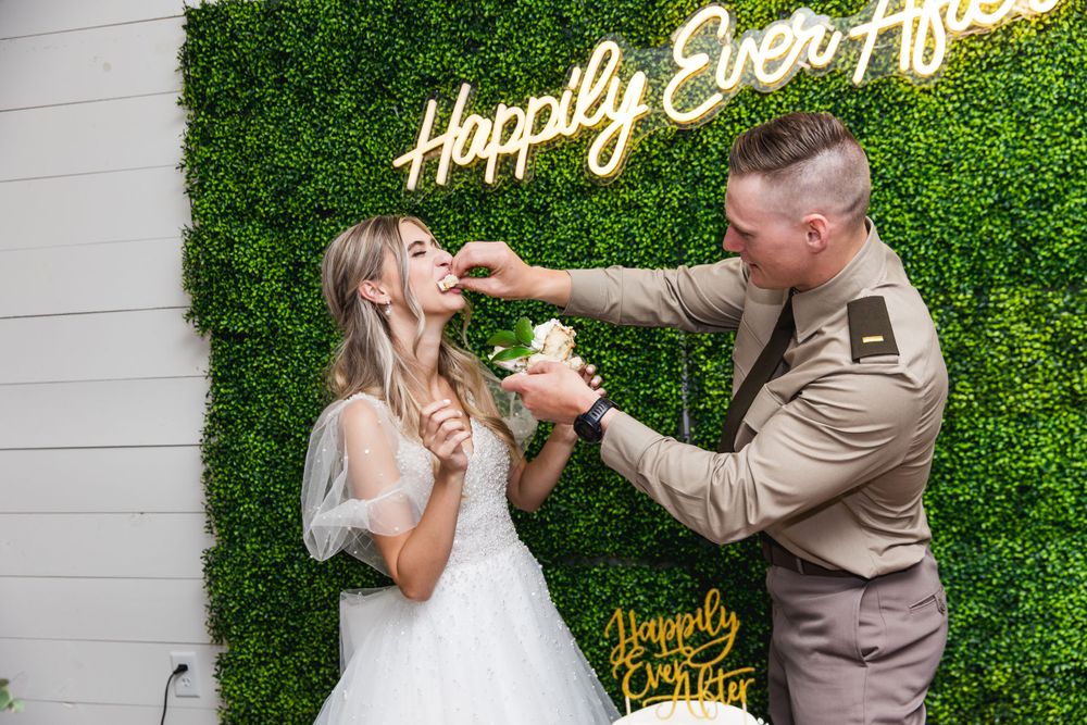 Newlyweds sharing cake at their wedding reception in front of a green hedge wall display.