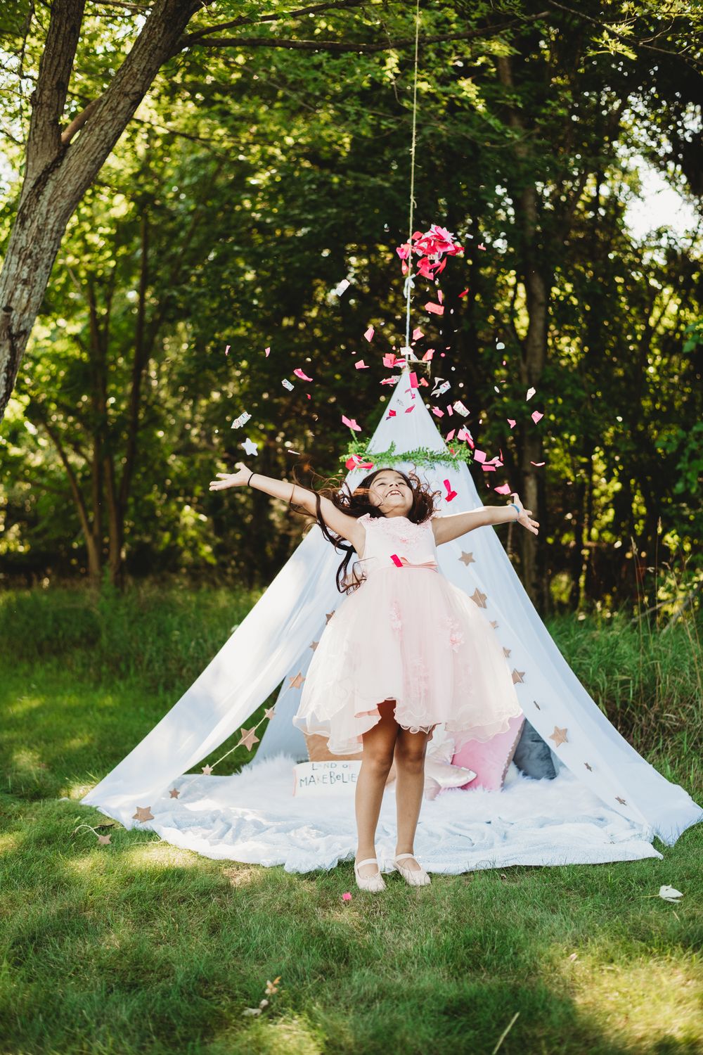 Whimsical outdoor scene with white teepee tent and pink flower petals scattered on grass.