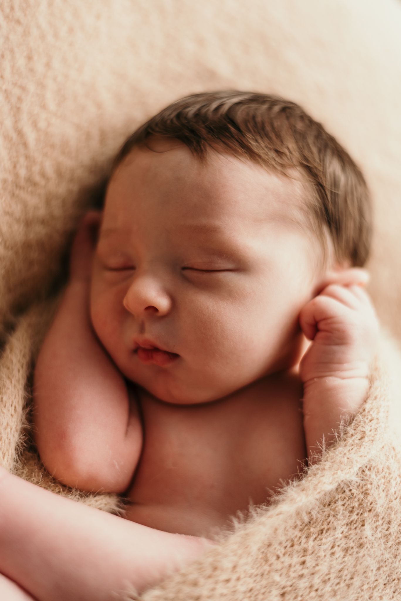 Sleeping newborn baby wrapped in cozy beige blanket during professional photoshoot.