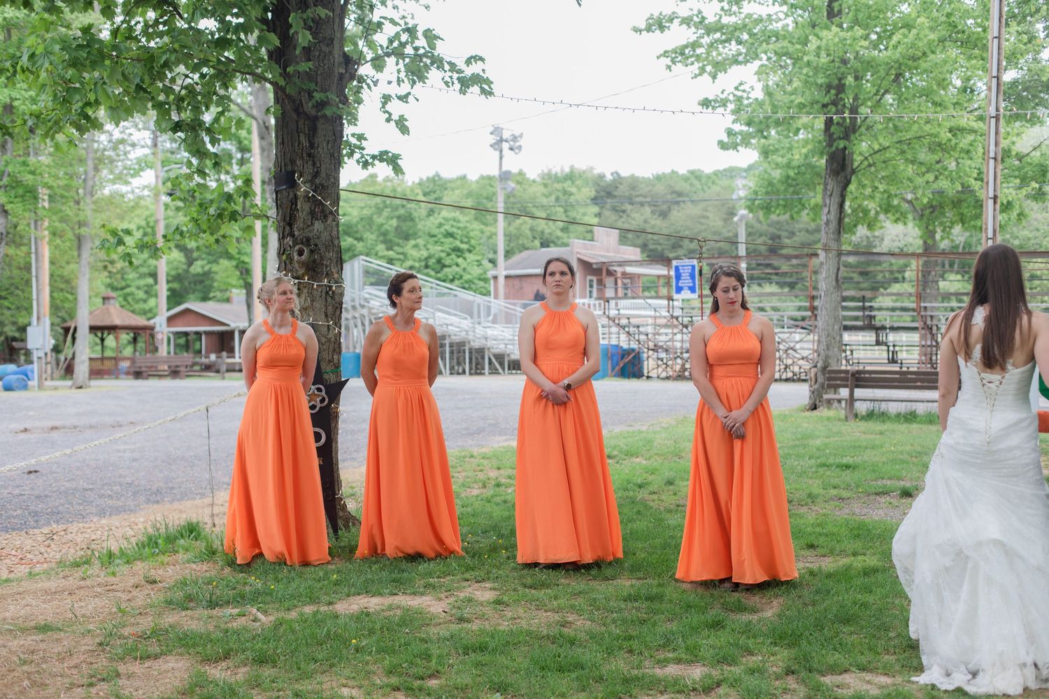 Four bridesmaids in matching orange floor-length dresses stand in a row on grass near trees.
