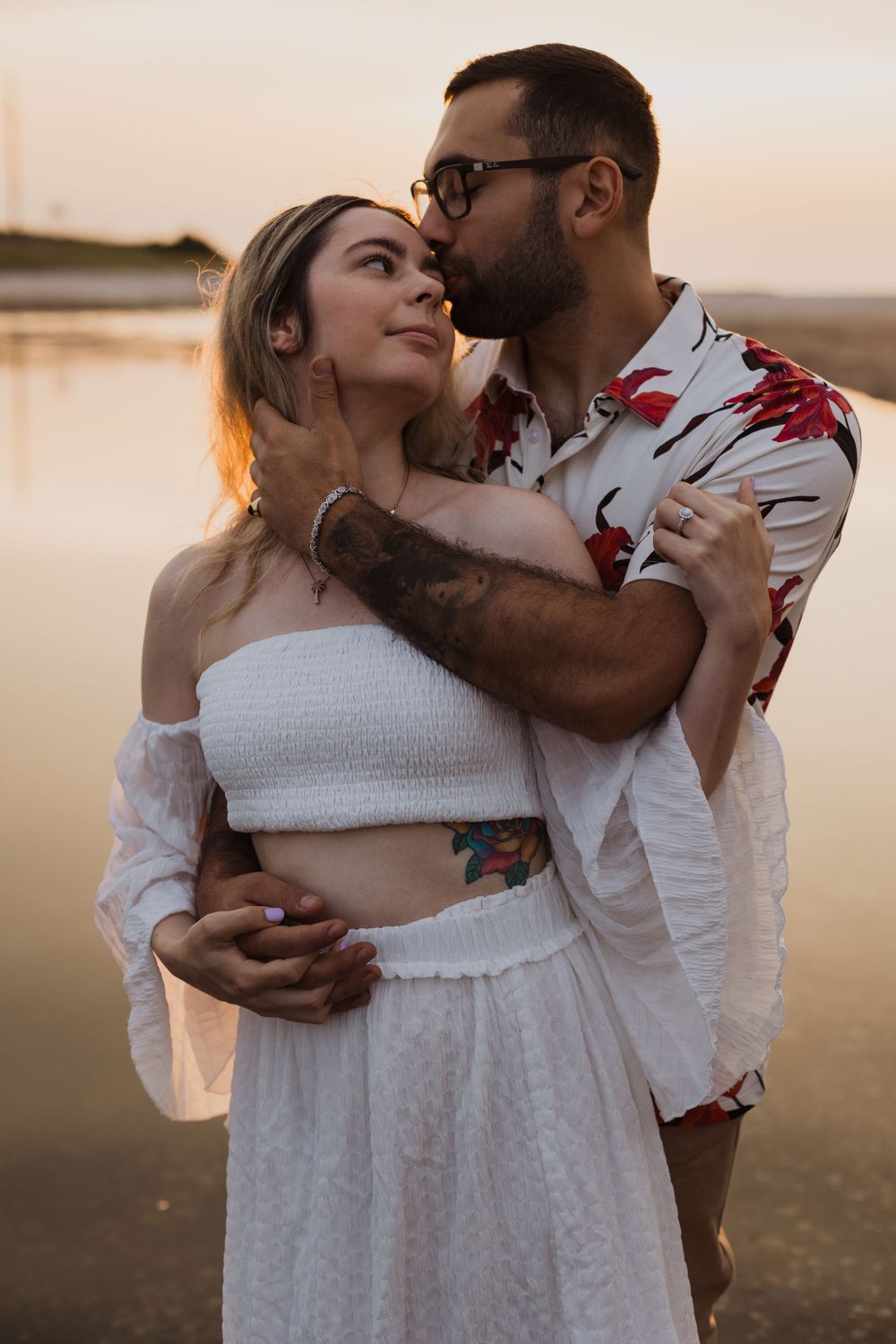 Intimate couple embrace on beach at golden hour wearing matching white and floral outfits.