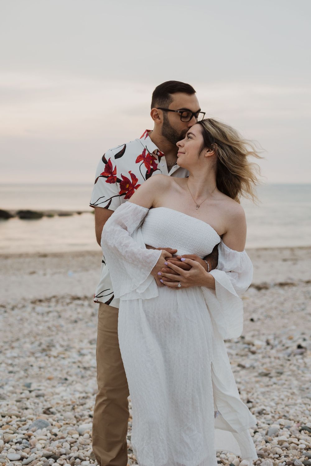 A romantic beach scene with two people embracing in white dress and floral shirt against cloudy sky.