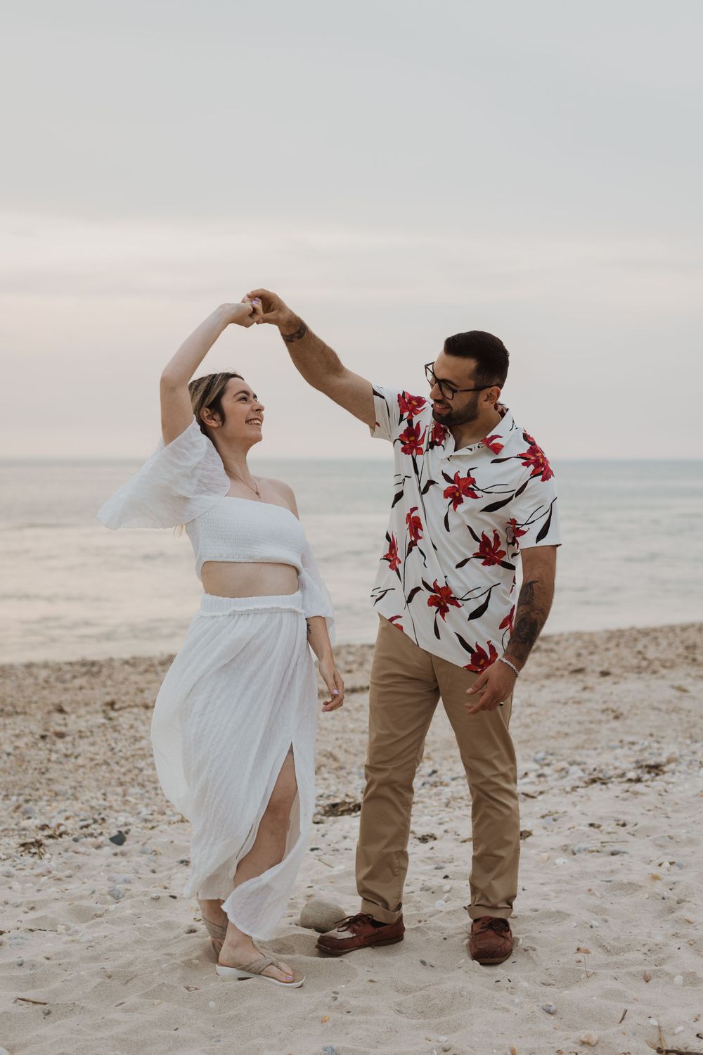 Couple dancing together on a beach at sunset wearing white dress and floral shirt.