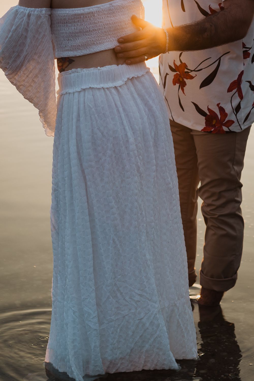 A close-up shot of embracing figures in white dress and floral shirt against sunset water.