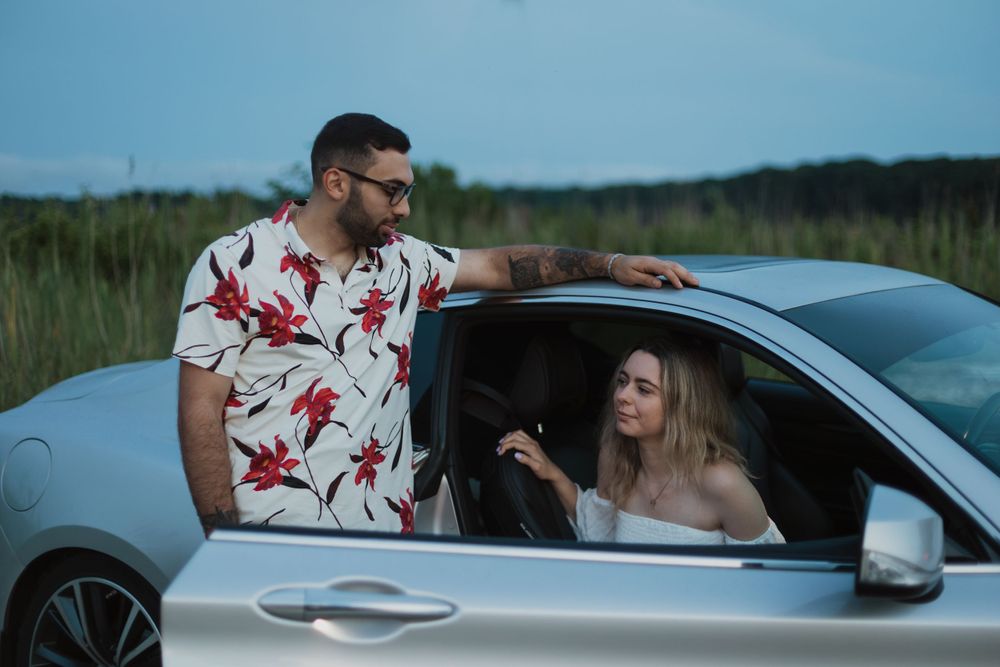 A couple shares a moment beside a silver car with natural scenery in the background.