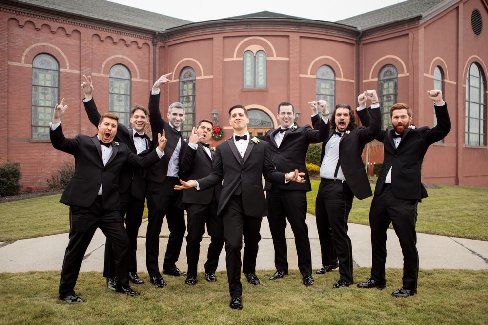 Groomsmen celebrate joyfully outside a brick church building in formal attire.