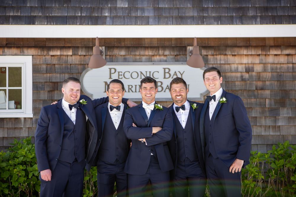 Groomsmen in navy suits pose outside the Peconic Bay building with cedar shingle siding.