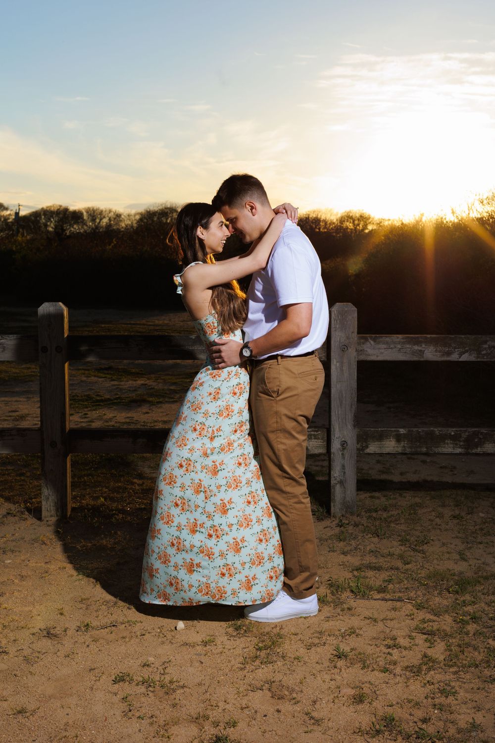 Couple embraces during sunset photo session by wooden fence in countryside setting.