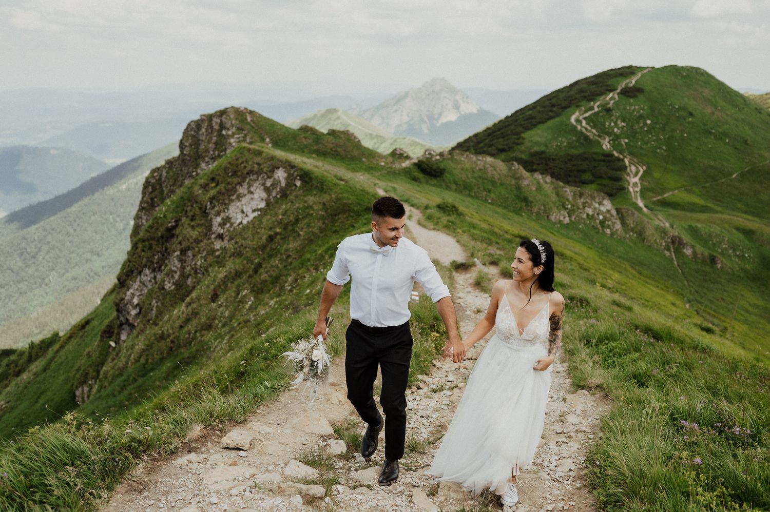 A couple runs joyfully along a mountain ridge trail in wedding attire.