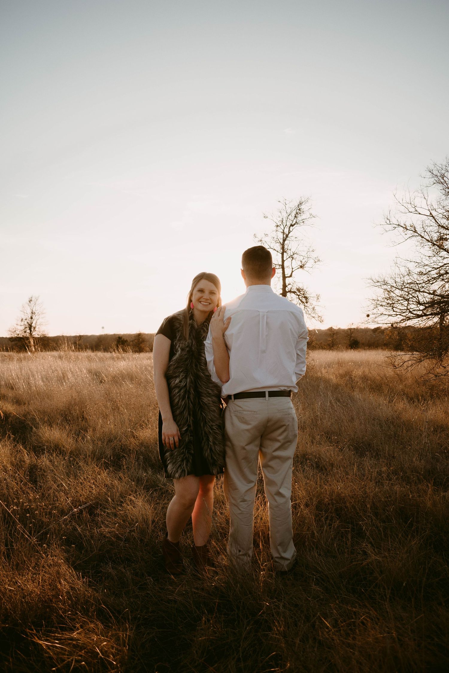 A couple stands together in tall grass during golden hour with bare trees in the distance.