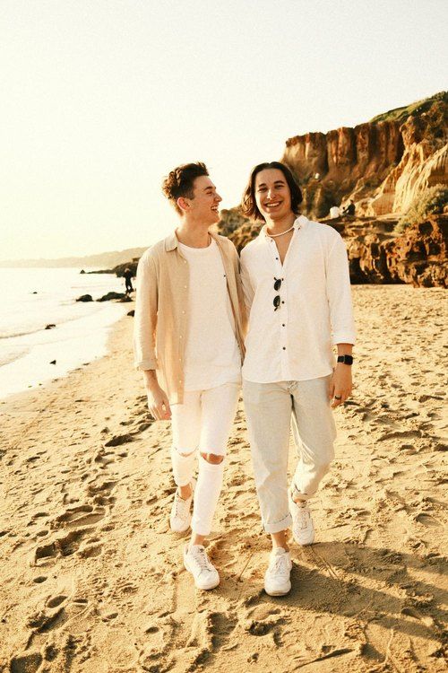 Two people in white outfits laughing together on beach with coastal cliffs in background.