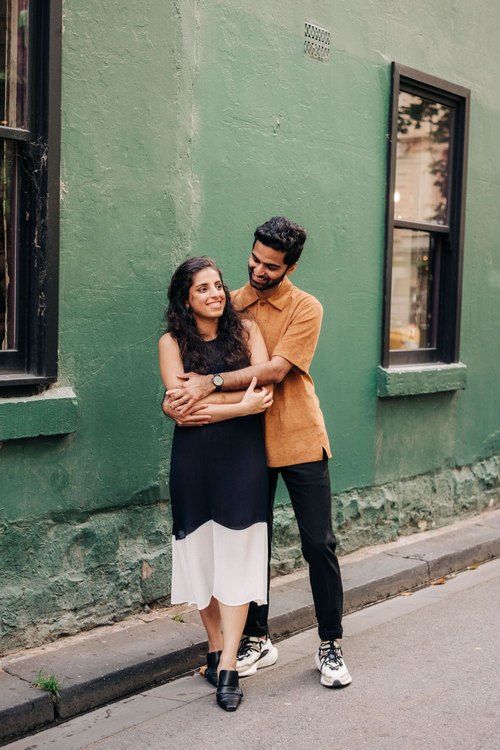 Couple embracing against green wall in urban setting with stylish casual outfits.