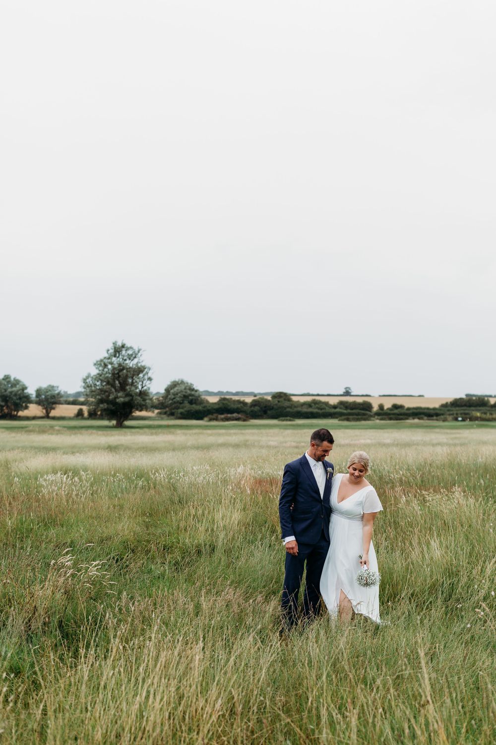 Couple walks through tall grass field on overcast wedding day.