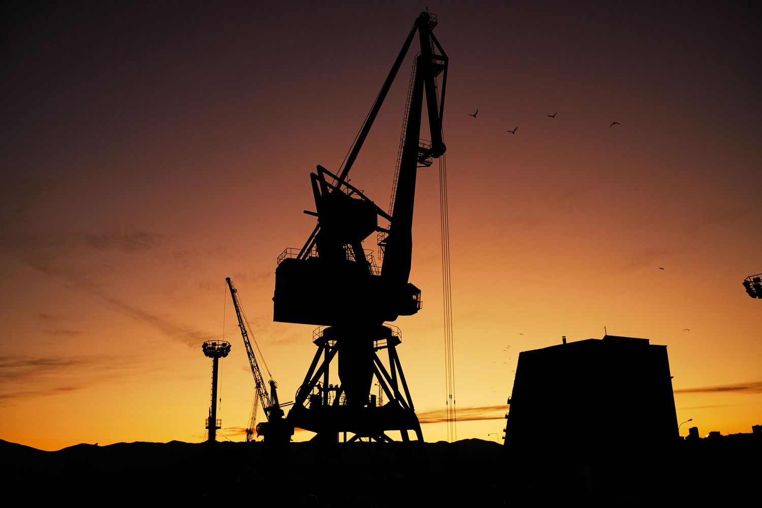 Silhouette of port cranes against dramatic orange sky at dusk.