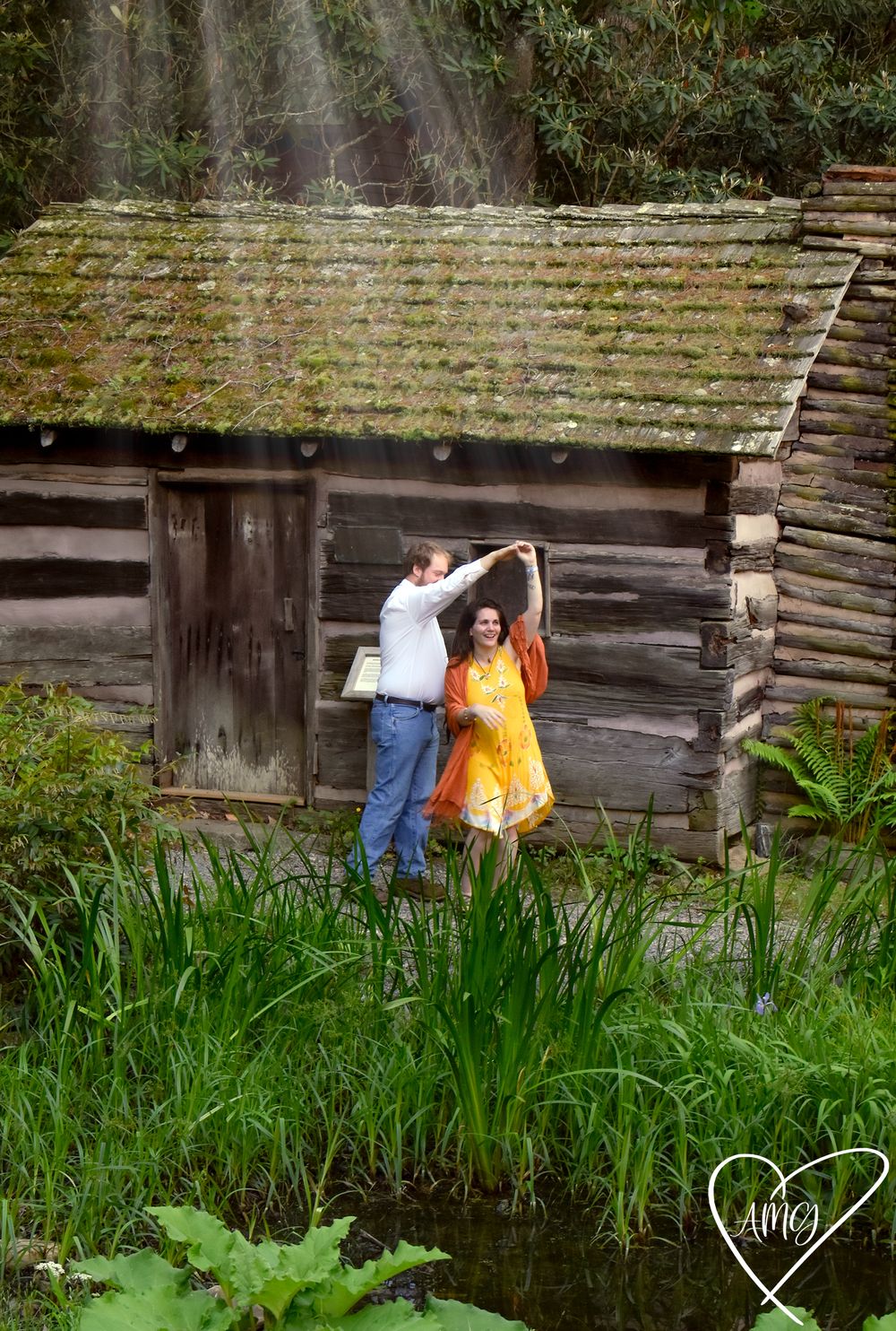 Couple dancing together in front of a rustic log cabin beside a pond, woman in yellow dress, lush green surroundings. Couples Photographer.