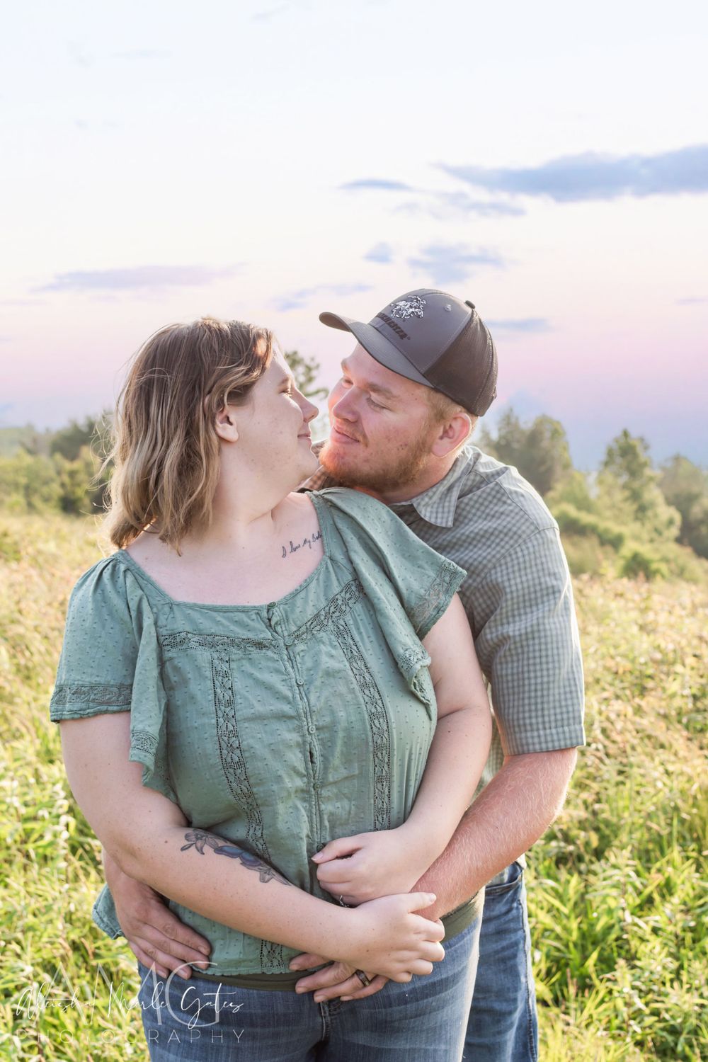 A couple shares a romantic moment in a sunlit meadow, the man nuzzling the woman in a green top. Maternity Announcement Photographer.