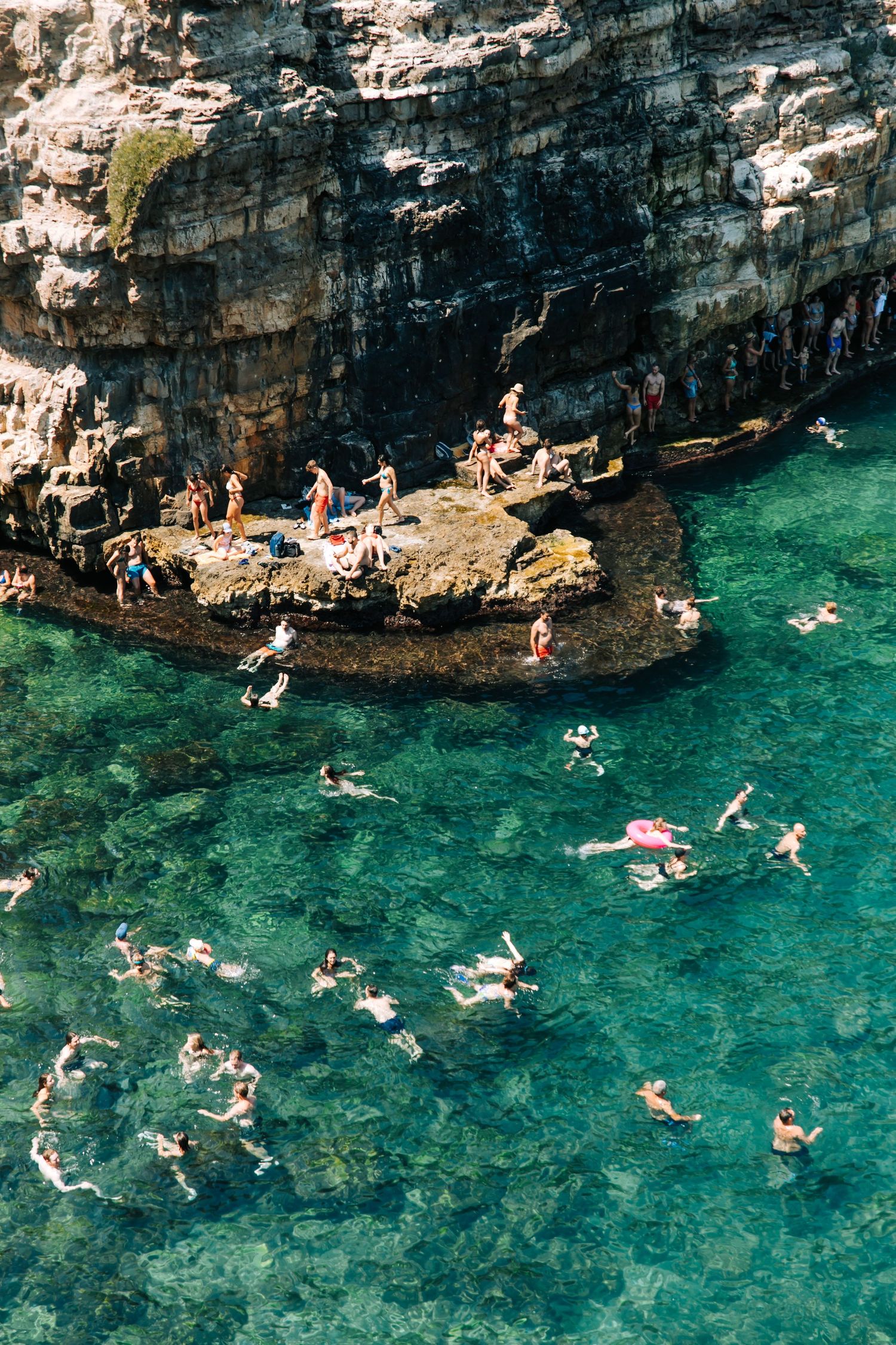 Aerial view of swimmers in crystal clear turquoise water beside rocky coastal cliffs.