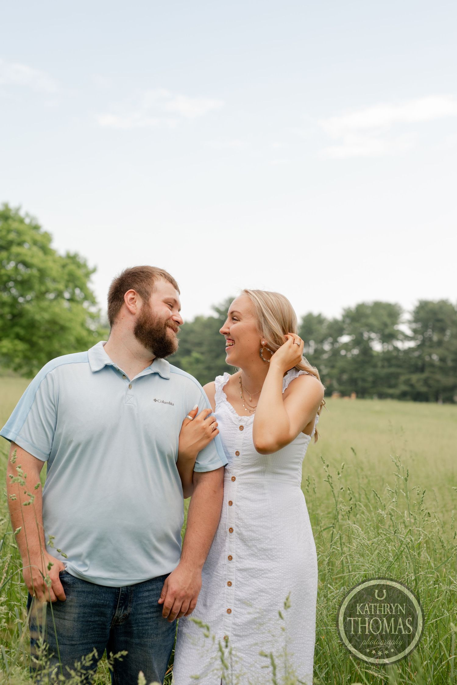 A couple shares a candid laughing moment together in a sunny meadow with tall grass and trees in the background.