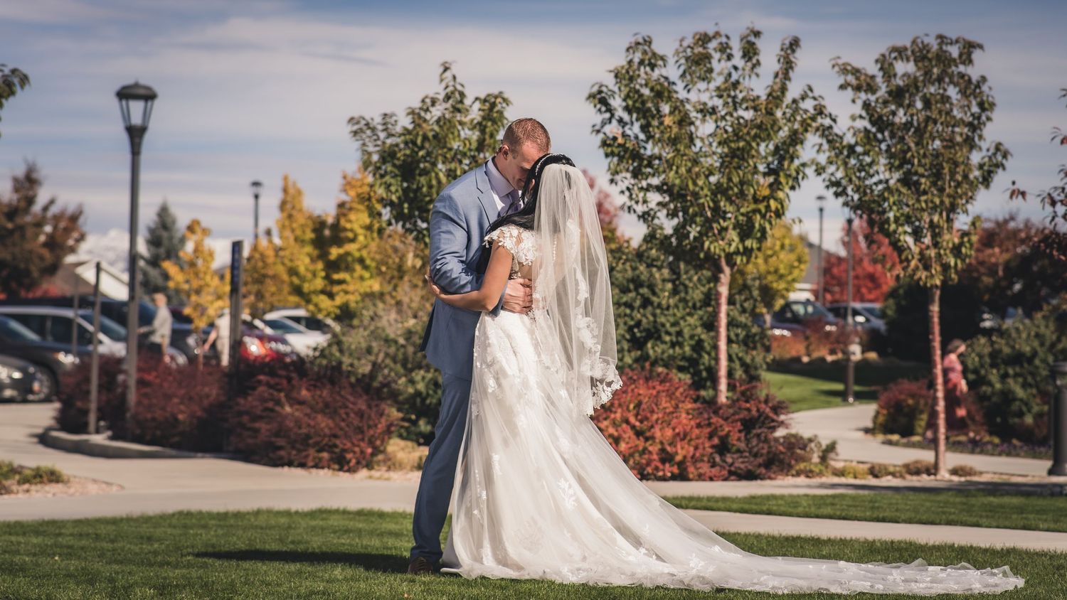 Bride and groom share a romantic kiss outdoors surrounded by colorful autumn trees.