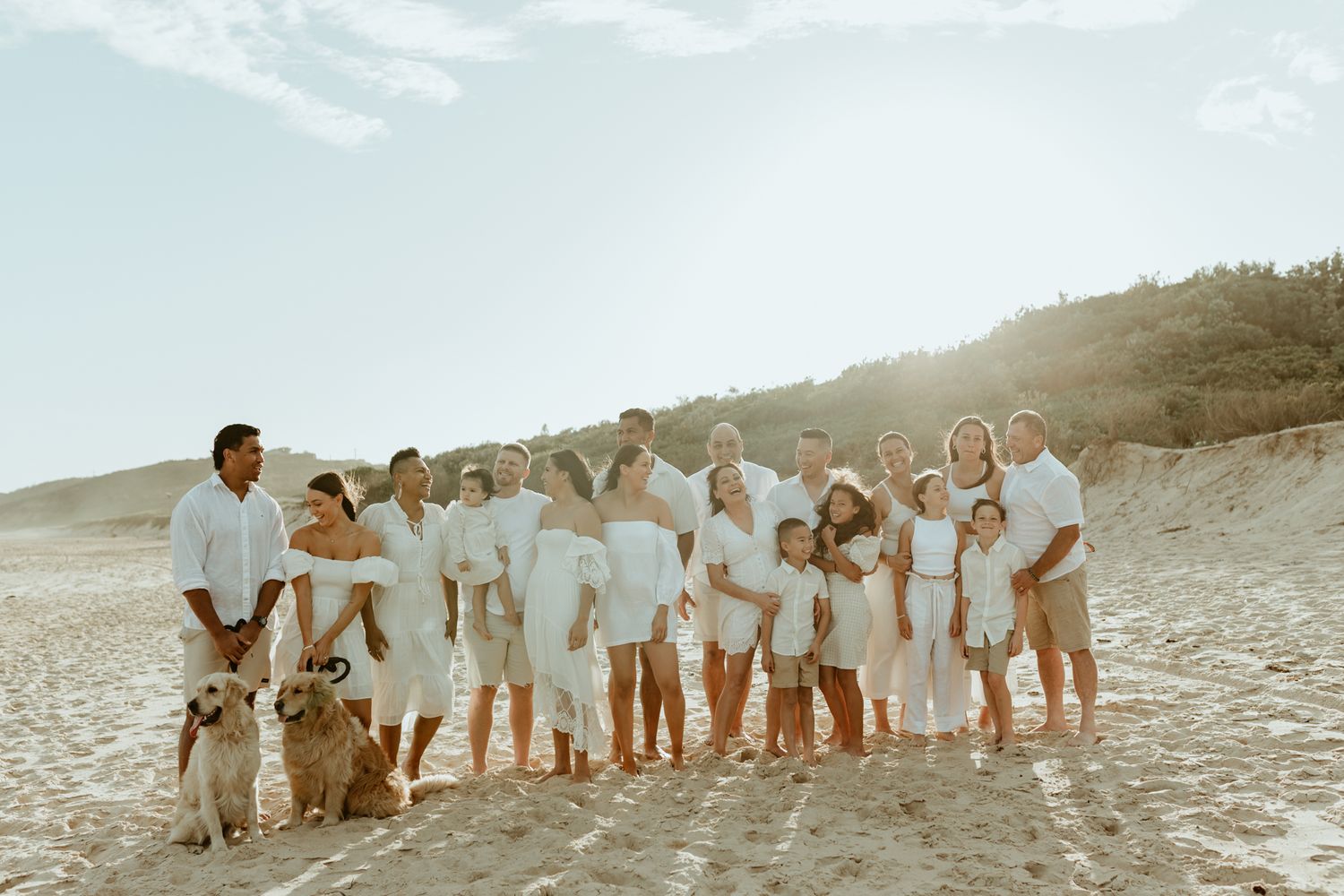 Large family group in white clothing poses together with dogs on a sunny beach.