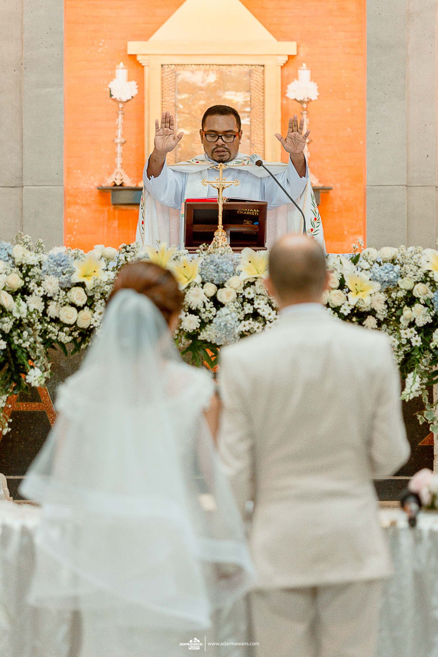 A religious ceremony takes place at an altar decorated with white flowers and orange lighting.