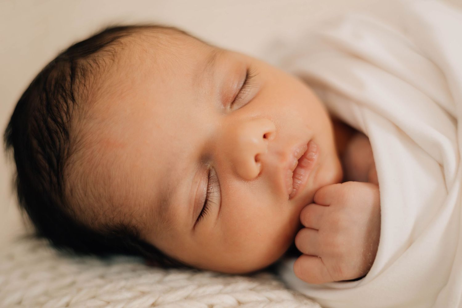 Peaceful sleeping newborn baby with dark hair wrapped in a white swaddle on a soft blanket.