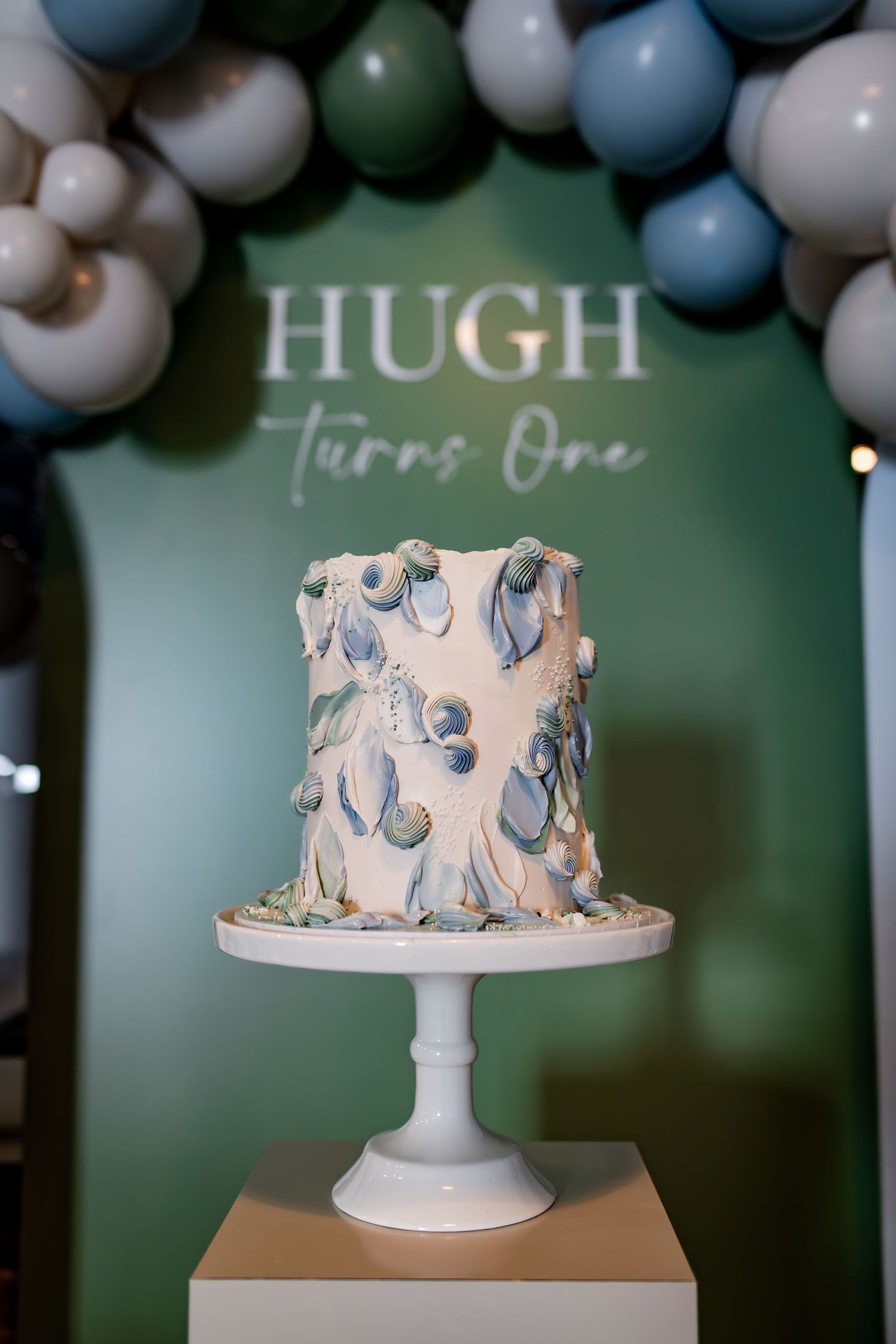 White and blue decorated birthday cake for Hugh featuring artistic swirl designs on a white pedestal stand.