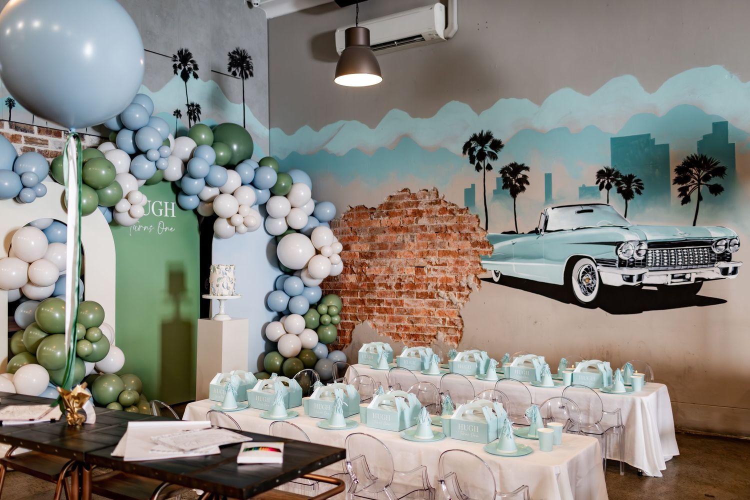 Retro-themed party space with balloon garlands and vintage car mural featuring palm trees and brick walls.