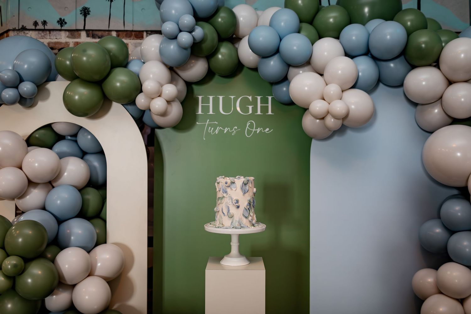 Party backdrop with balloon arch and decorative cake display reading 'Hugh Turns One'.