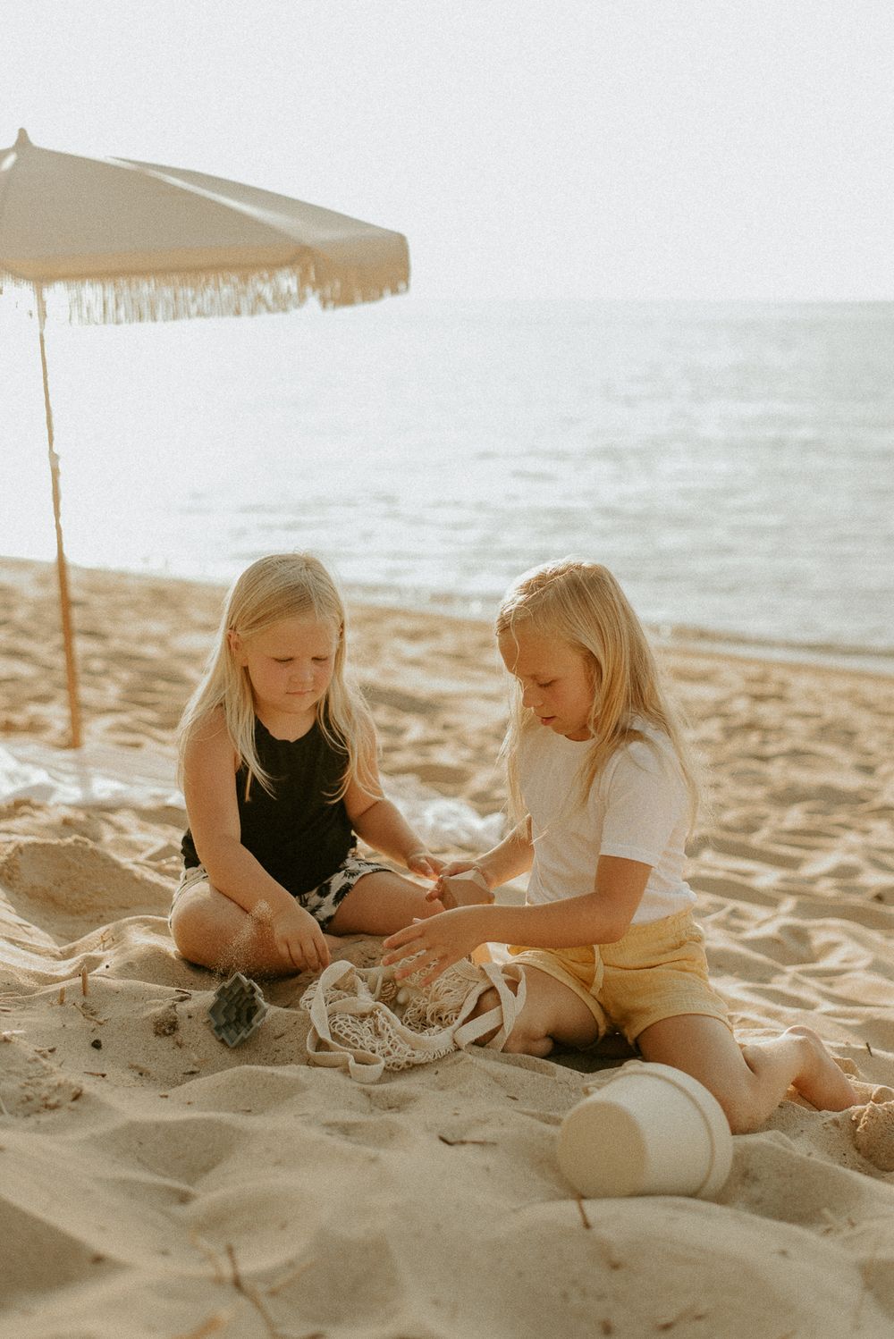 Two children playing with sand toys under beach umbrella by ocean.