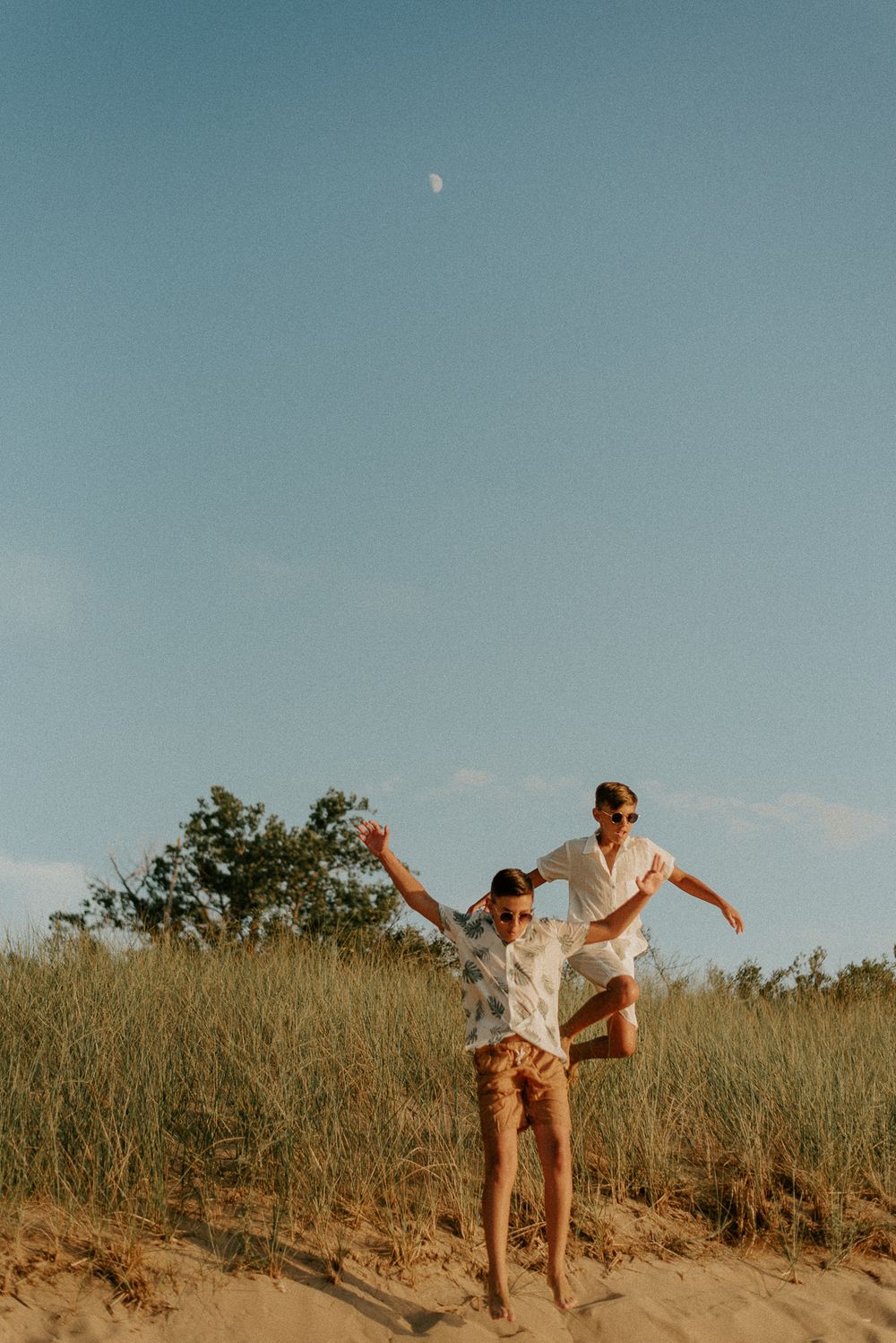 Two people playfully pose on a sandy dune with the moon visible in the blue sky.