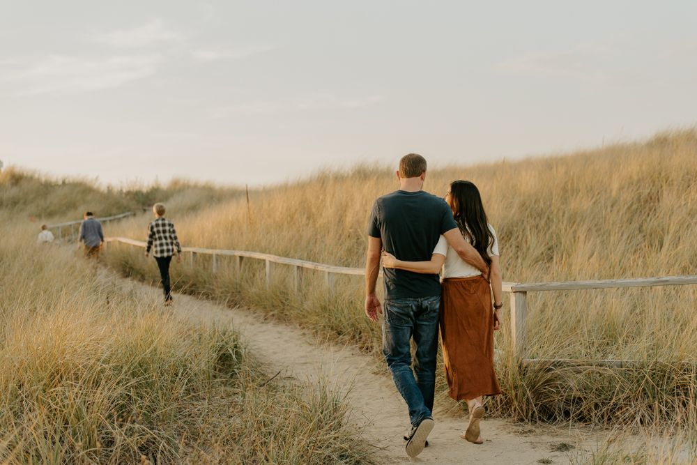 Couples walk together on a winding boardwalk path through tall grass at sunset.