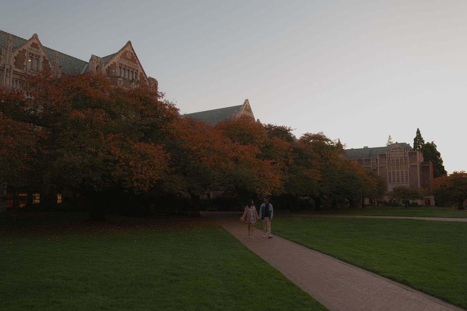 A stone building with Gothic architecture sits on a grassy lawn at dusk with people walking on a path.