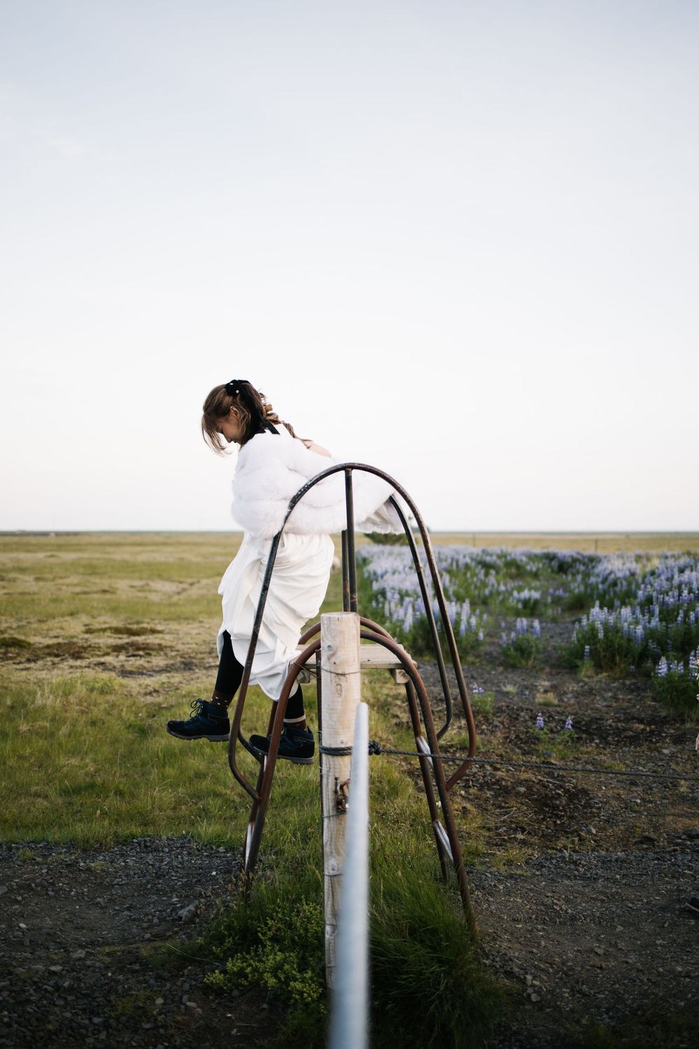 Someone in white wedding attire balances on metal railings in an open field at dusk.